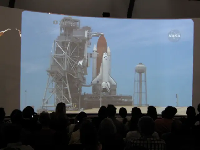In this image, we can see a crowd in front of the screen. On the screen, there is a rocket...