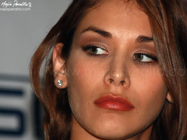 In this image there is a face of a woman. She is wearing earrings. The background is blurry. In the...