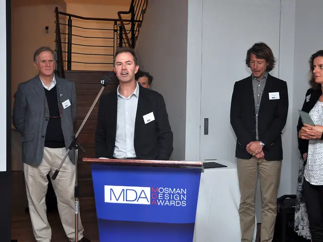 In this image there are people standing and a man standing near a podium and there is a mike, in...