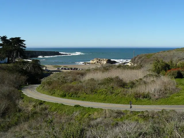 In this picture we can see ocean. On the bottom we can see road, grass, plants. On the left we can...